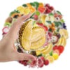 A hand holds a transparent sticker featuring sliced yellow fruit on a background of various colorful berries and fruits.