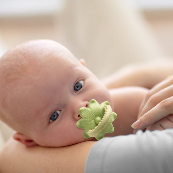 A close-up of a baby's face with a silicone teething toy shaped like a green flower and grape resting near the mouth.