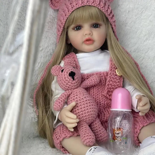Muñeca bebé con largo cabello rubio, vistiendo un atuendo de punto rosa y una botella rosa.