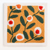 A square piece of felt featuring an orange background with green leaves and white and orange flower embroidery.