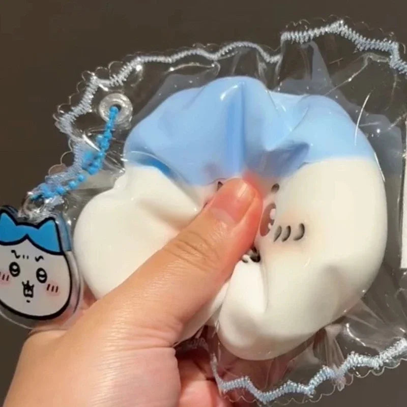 A hand is holding a squishy stress relief toy shaped like a white and light blue face inside clear plastic packaging.