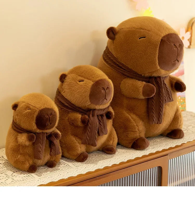 A brown plush capybara stuffed animal wearing a knit scarf sitting on a wooden surface.