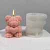 A pink silicone candle mold shaped like a bear hugging a heart, next to a lit candle.