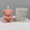 A pink silicone candle mold shaped like a bear hug, next to a lit candle.
