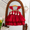 A red velvet dress for a baby girl featuring ruffled sleeves and pink pom-pom details displayed in a wooden mirror frame.