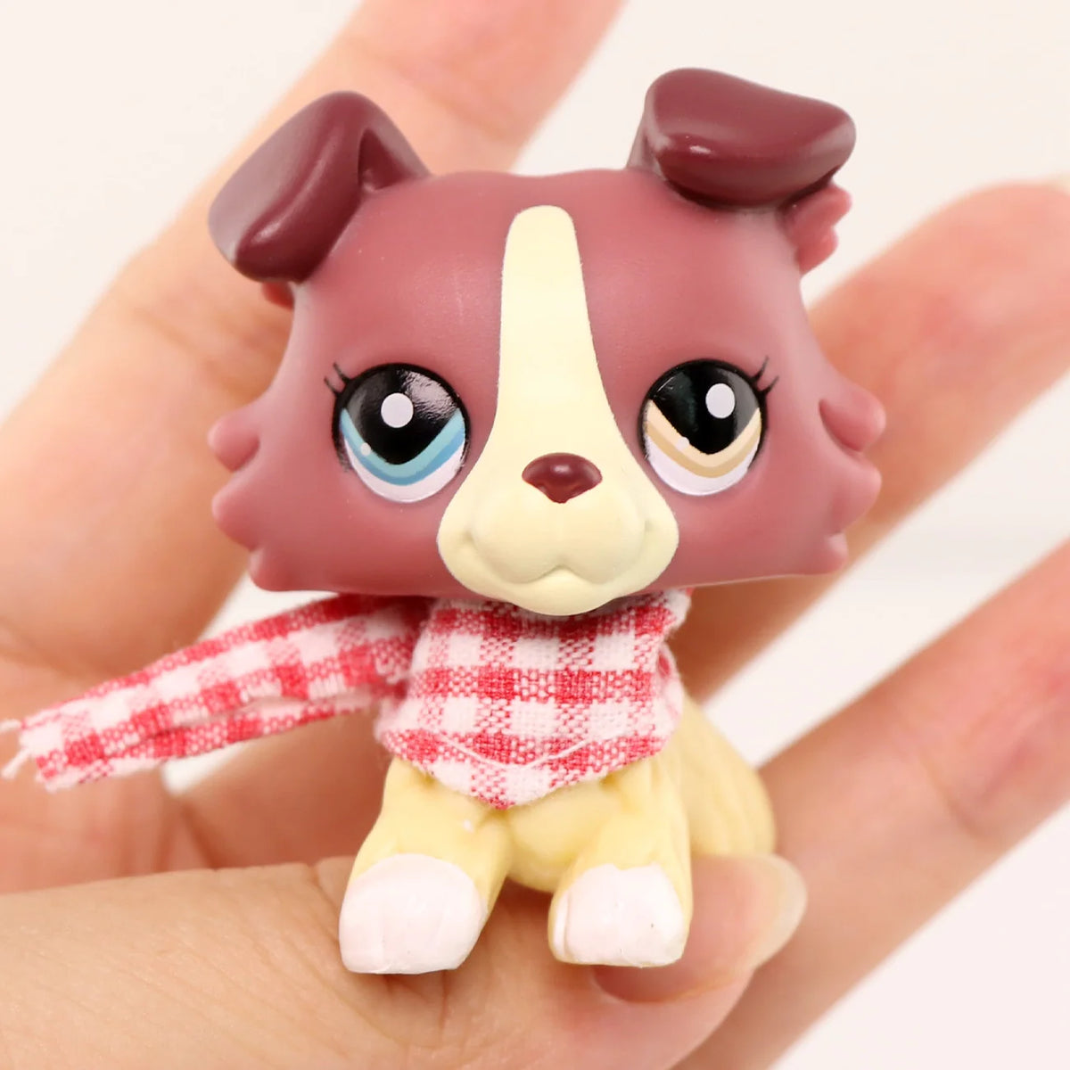 A small, brown and white miniature pet animal figure with a red and white checkered shirt accessory is held in a hand.