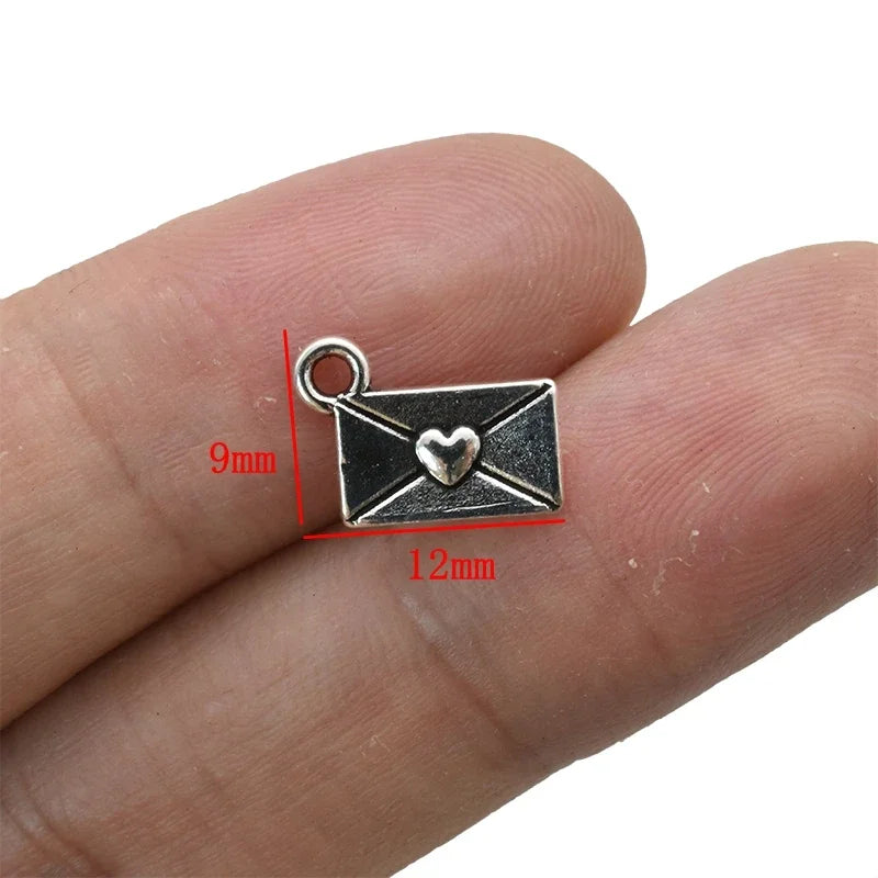 A silver vintage envelope charm with a heart design is shown resting on a human finger.