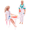 A blonde doll dressed as a doctor wearing a white lab coat and stethoscope stands next to a white table with medical accessor