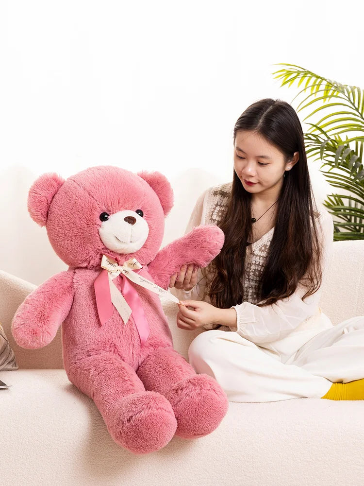 A young woman is sitting on a couch holding a large pink teddy bear plush toy.