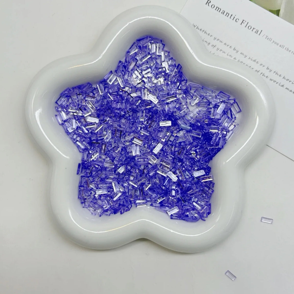 A white flower-shaped dish filled with purple iridescent acrylic ice strips for nail art and DIY crafts