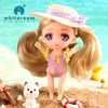 A small doll with long blonde hair wearing a purple outfit and a straw hat standing on a sandy beach.