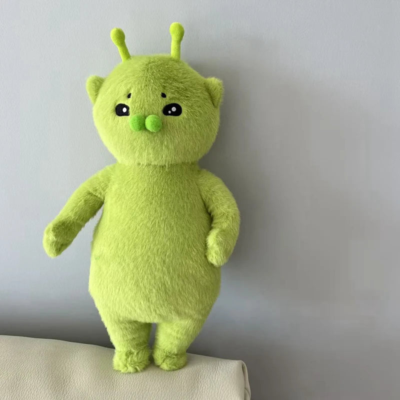 A bright lime green alien cat plush toy with large eyes and green nose standing against a light gray wall.