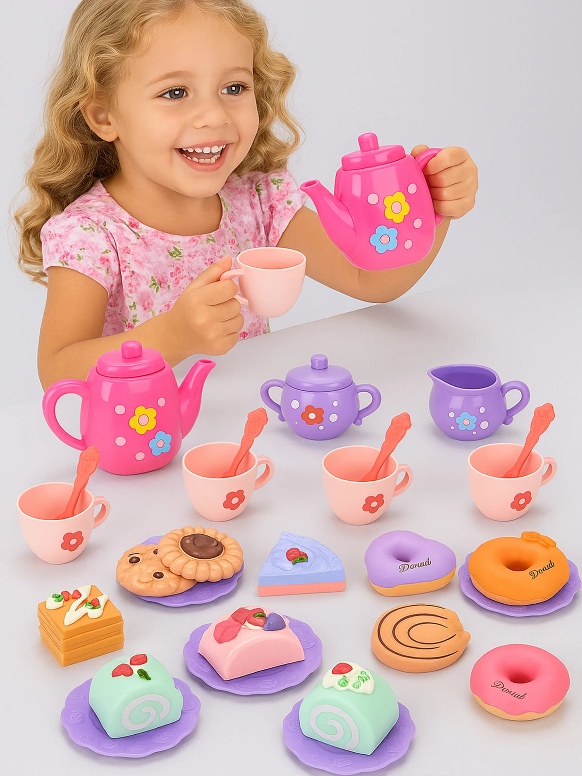 A young girl is holding a pink teapot and pouring tea into cups, surrounded by various colorful pretend play food items.