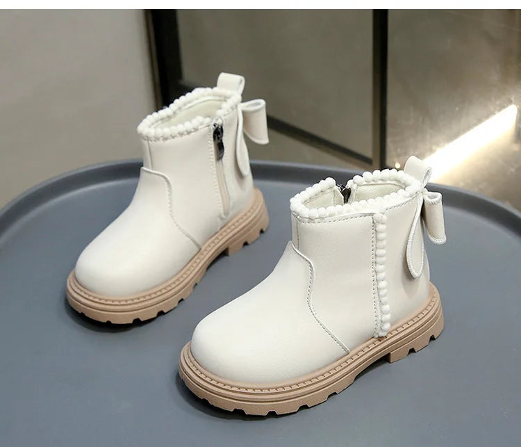 A pair of white faux leather winter boots with white fluffy trim and light tan soles displayed on a gray surface.