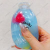 A hand holds a clear blue squishy fidget toy bottle with a pink fruit design.