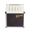 A white guitar pick holder with a speaker-shaped design featuring dark brown stripes and gold lettering.