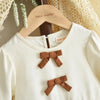 A white girls' long sleeve dress with brown bow details displayed on a wooden hanger.