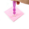 A hand holds a purple diamond painting clay stick resting on a pink square surface.