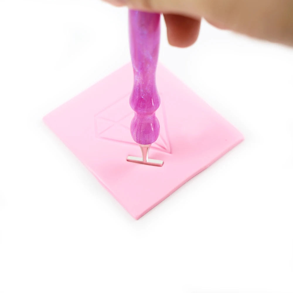 A hand holds a purple diamond painting clay stick resting on a pink square surface.