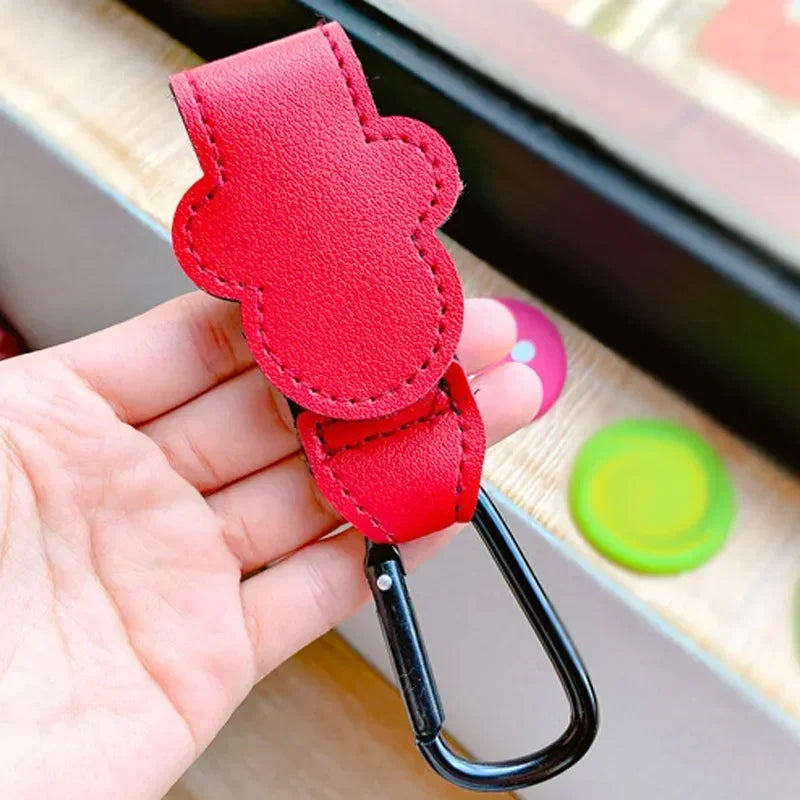 A hand holds a red PU leather stroller organizer hook with a black clip against a light background.