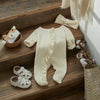 A cream knit ruffle romper for a baby girl lying on wooden stairs next to a headband and shoes.
