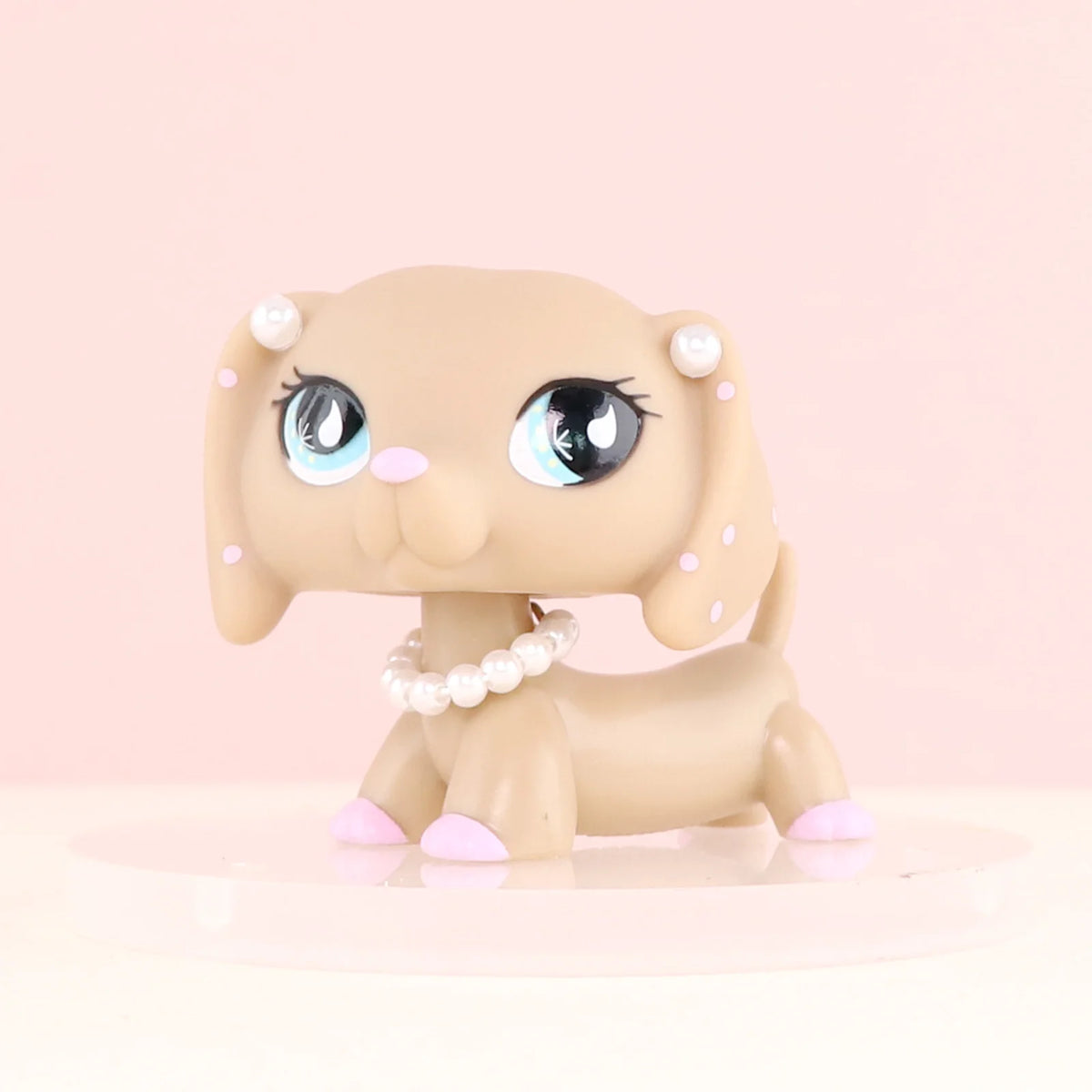 A light beige miniature dog figure with large blue eyes and pearl accessories sitting on a pink surface.