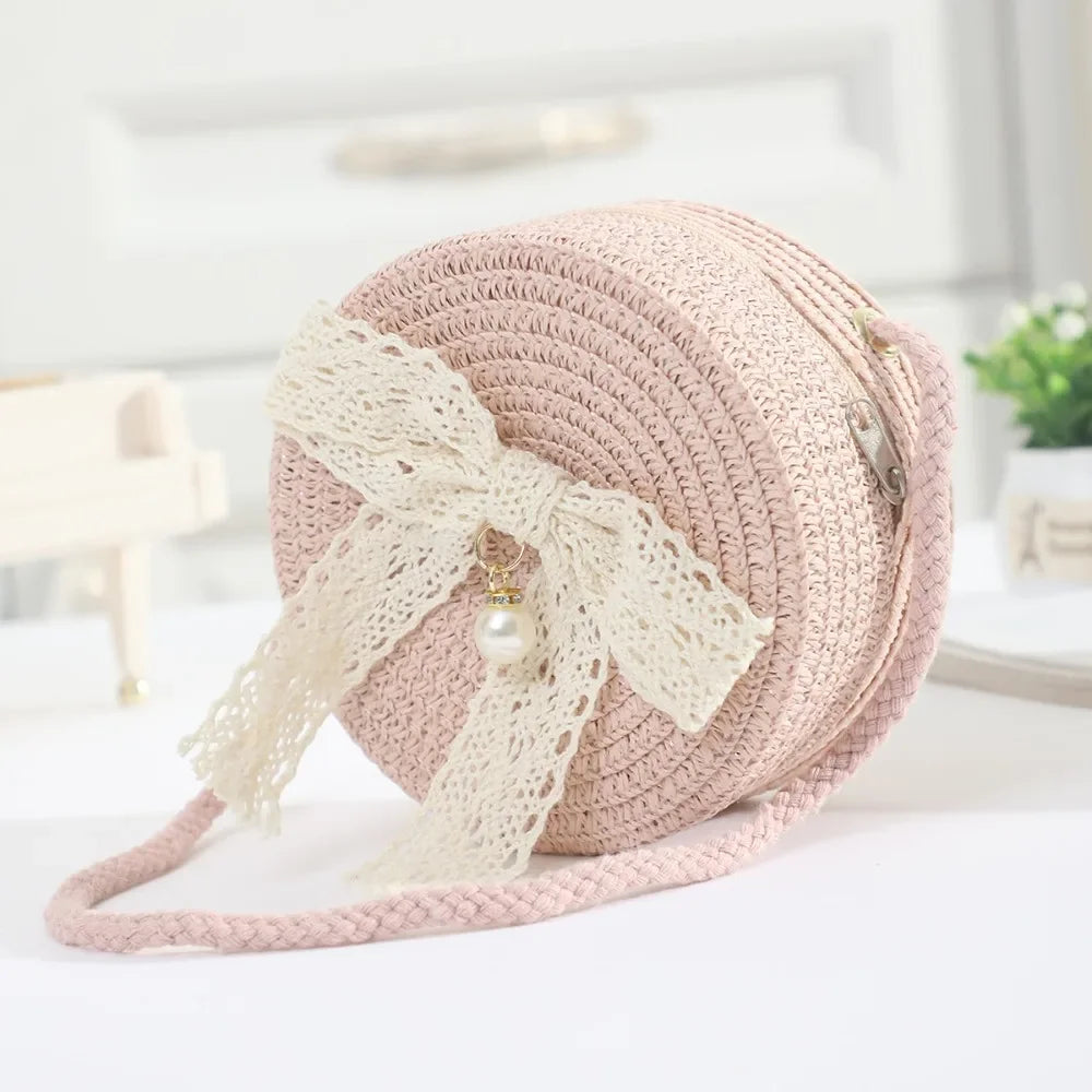 A light pink handwoven straw crossbody bag featuring a delicate white lace bow detail.