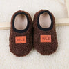 Pair of brown fuzzy baby slippers with black lining and orange WLS labels resting on a white fluffy surface.