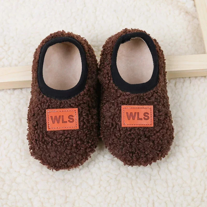 Pair of brown fuzzy baby slippers with black lining and orange WLS labels resting on a white fluffy surface.