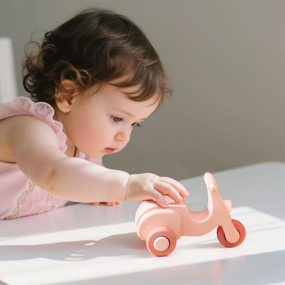 A baby is interacting with a pink silicone toy car set on a white surface.