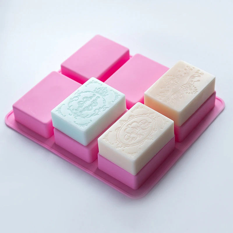 A pink silicone mold tray holding six square bars of handmade soap with floral patterns.