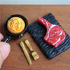 A miniature dollhouse set featuring a red steak and pasta food item served on a black textured plate with small gold cutlery.