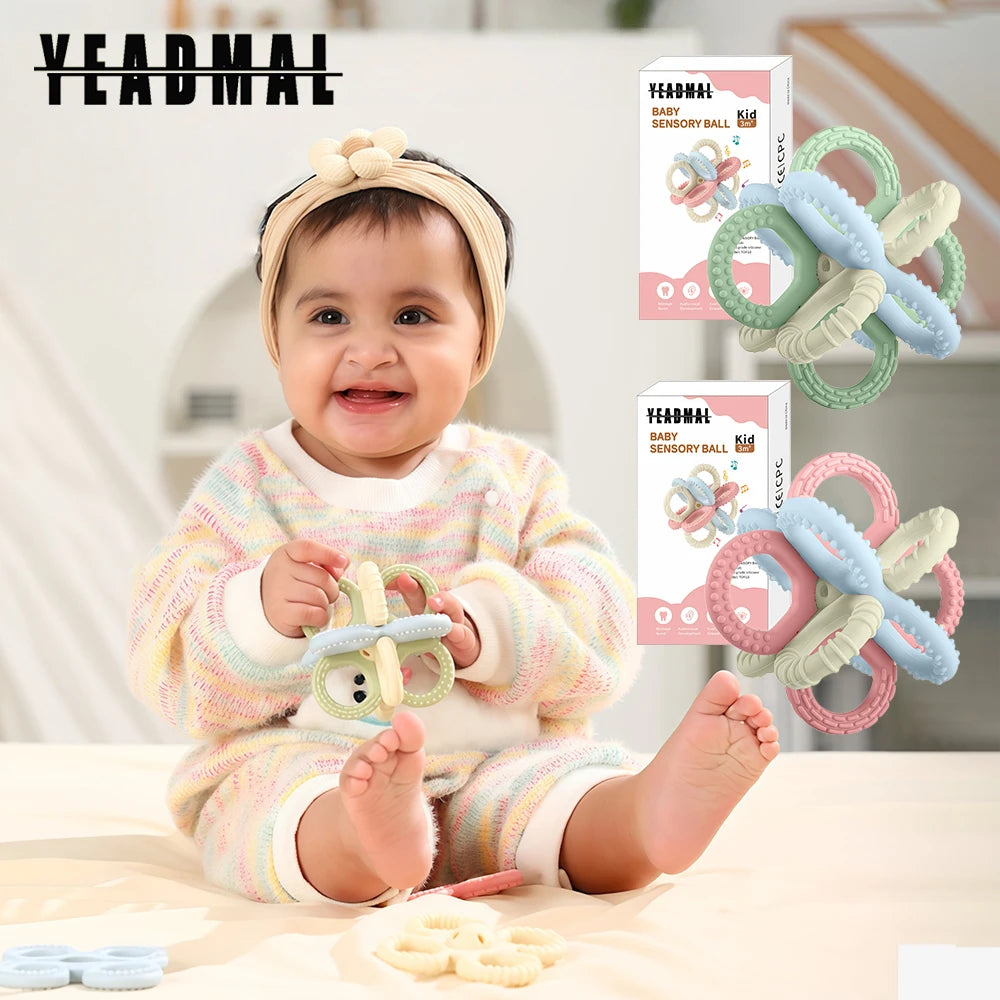 A baby is playing with a set of silicone teether toys, including rings and rattles, on a light wooden surface.