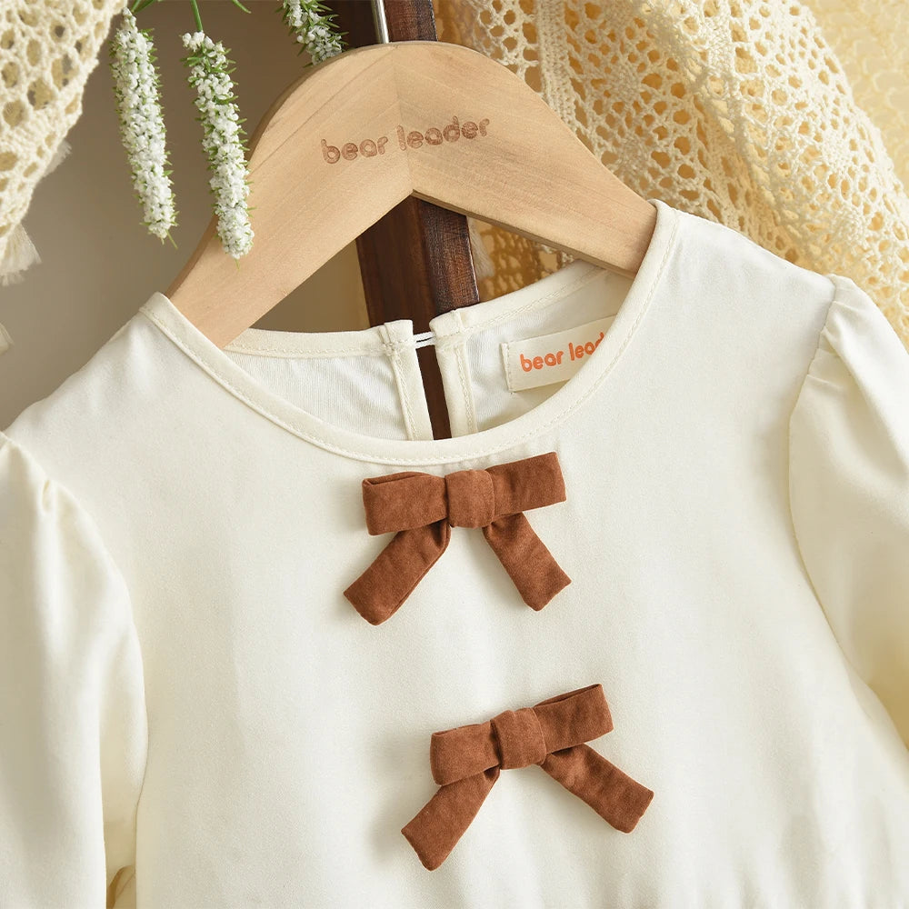 A cream long sleeve dress with brown bow details displayed on a wooden hanger.