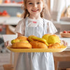 A young girl in an apron holds a tray of pretend play food items like bread rolls and pastries.