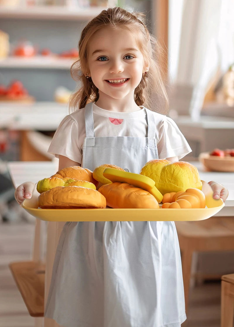 A young girl in an apron holds a tray of pretend play food items like bread rolls and pastries.