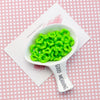 A white spoon holds bright green, wavy acrylic beads with a floral edge on a pink background.