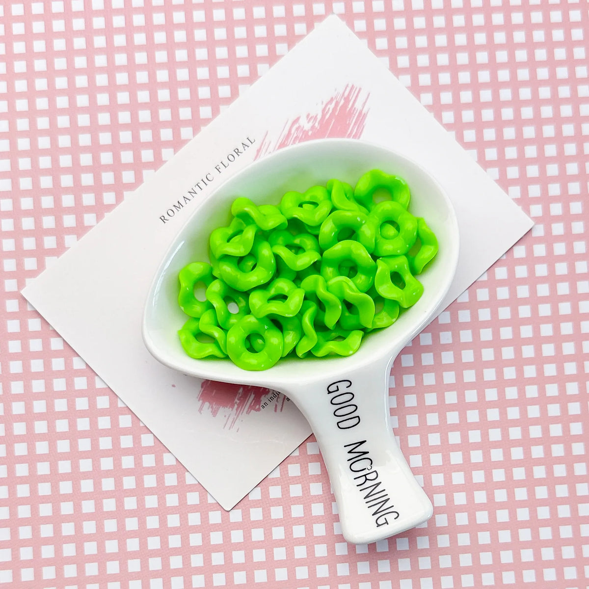 A white spoon holds bright green, wavy acrylic beads with a floral edge on a pink background.