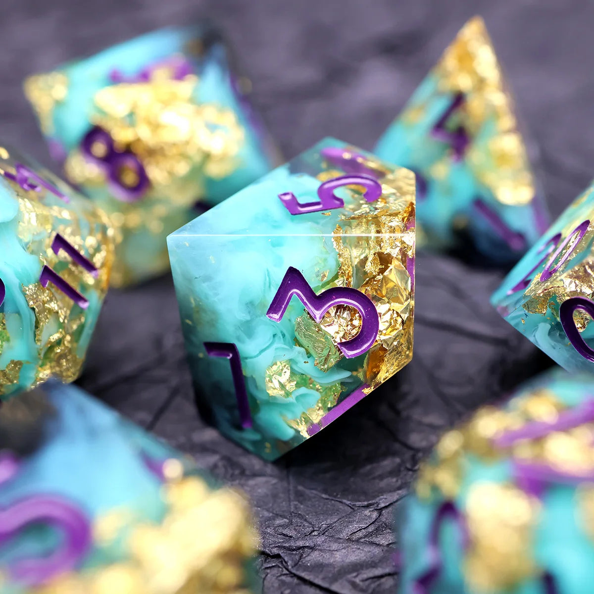 A close-up of several teal resin polyhedral dice with gold accents scattered on a dark surface.