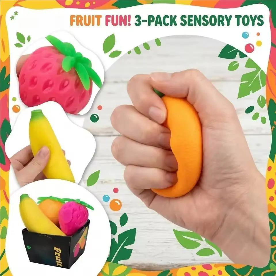 A hand holds a set of three fruit sensory toys including a pink strawberry, yellow banana, and orange, against a green leafy 