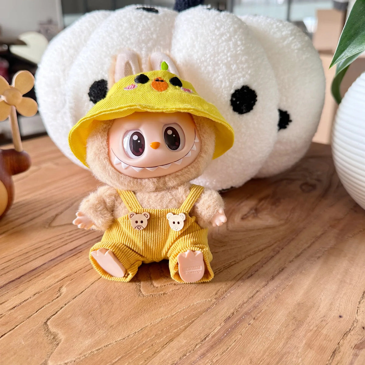 A yellow Labubu doll wearing a yellow hat and overalls, set against a white polka-dotted background.