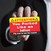 A hand holds up a yellow warning card with text that reads ATTENTION You Parked Like an Idiot Flip over to see the reason