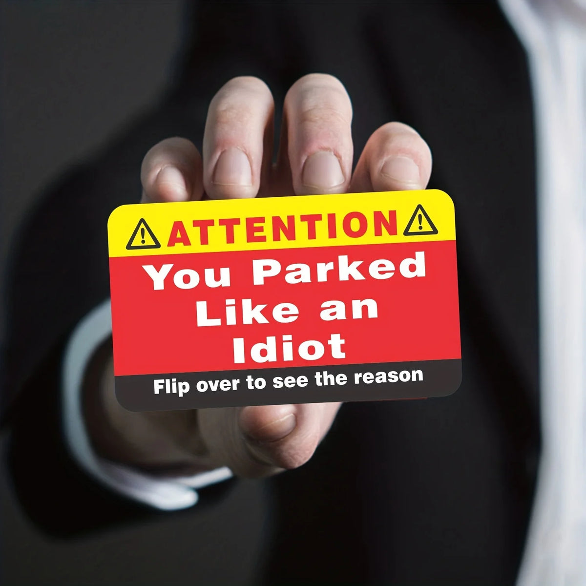 A hand holds up a yellow warning card with text that reads ATTENTION You Parked Like an Idiot Flip over to see the reason