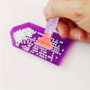 A hand is using a plastic tool to apply white beads onto a purple diamond painting tray.