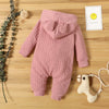 A pink knitted hooded baby romper lying on a wooden surface next to a small yellow smiley card and some greenery.