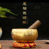 A golden copper singing bowl with a wooden mallet resting on a red and gold fabric surface.
