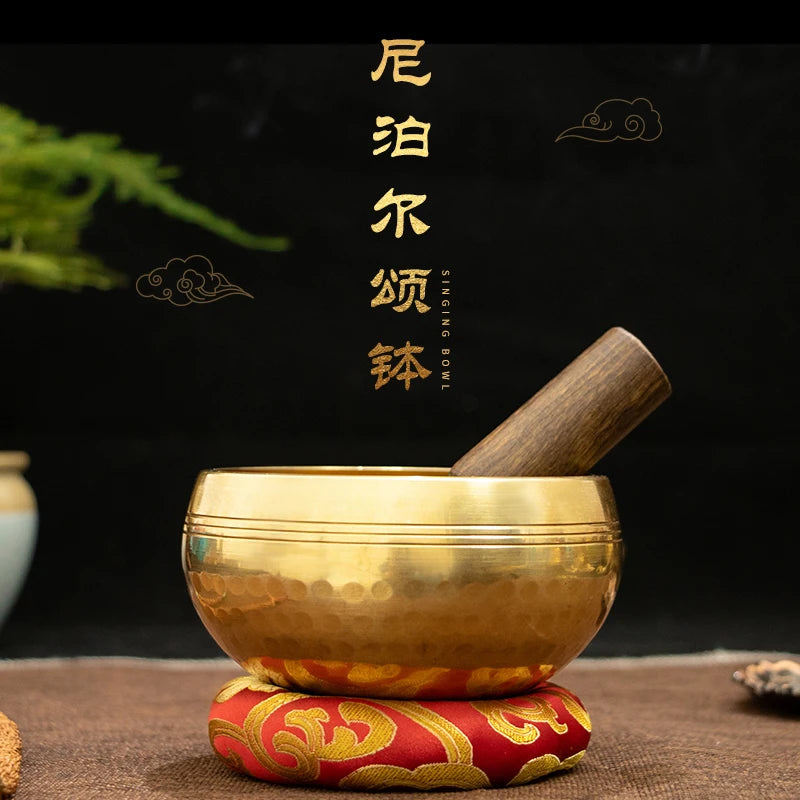 A golden copper singing bowl with a wooden mallet resting on a red and gold fabric surface.