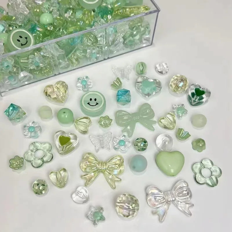 A collection of clear and green acrylic bow and flower beads for jewelry making displayed in a clear container.
