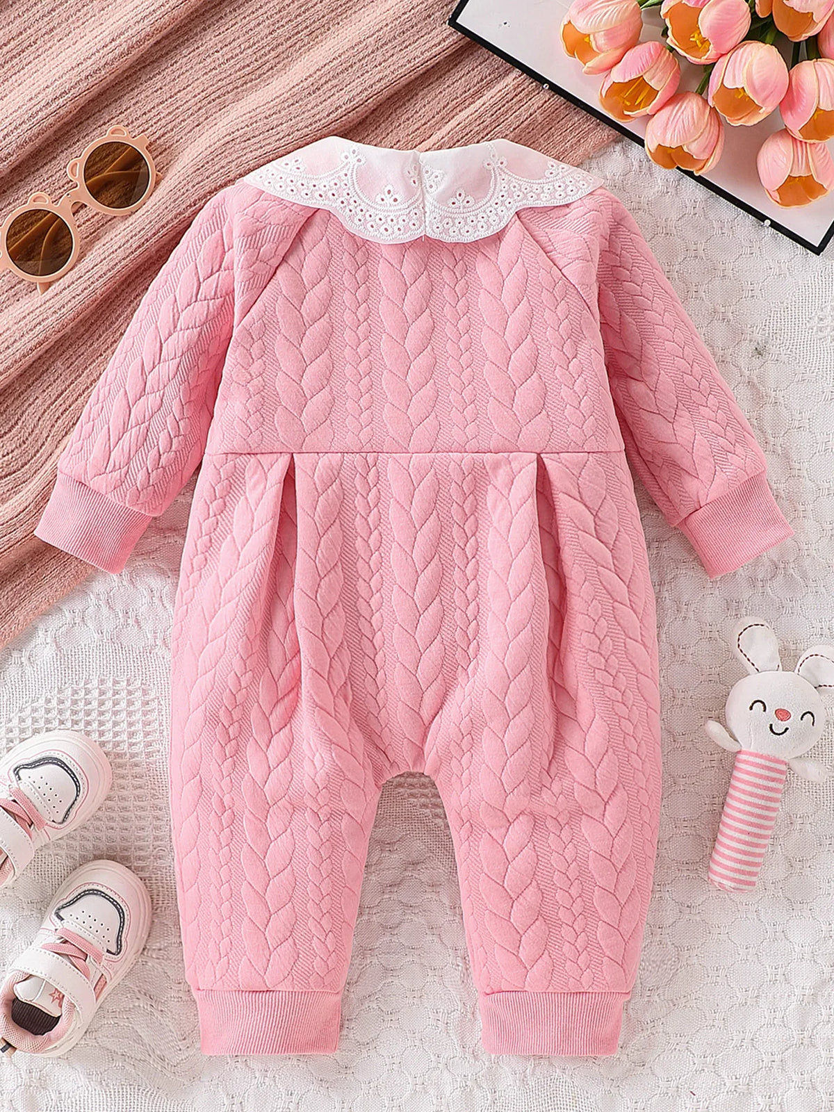 A pink quilted baby girl romper with a white lace collar lying on a white textured surface.