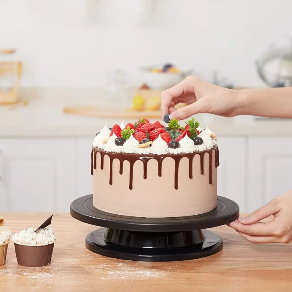 A hand is decorating a round cake with frosting and fruit using tools on a wooden table.